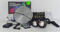 Photo of the full set including the waterproof LED strip light coiled, the remote control, power adapter, two landscape spotlights with stakes, instruction manual, and original box.
