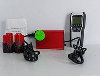 Full set showing two rechargeable lithium batteries, red battery charger with power cords, digital voltage tester, and charger instruction manual arranged on flat surface.