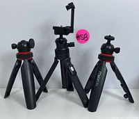Three black adjustable tripods including one with smartphone clamp and two with ball head mounts with red rings, shown front view.
