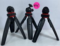 Three Mobifoto tripods shown from the front with flexible legs, black and red design, and rotating ball heads. One tripod has a visible remote shutter button on the handle.