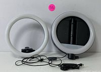 Two round LED ring lights shown from front with white light rings and black centers, one with stable base and control switch.