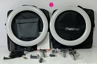Front view of both Mobifoto LED ring lights placed on black soft carrying cases along with remote control, power adapters, mounting accessories, and gray fabric pouches for accessories.
