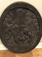 Close-up front view of an oval metal relief showing four cherubs in a classical scene among trees with a dark patina finish.