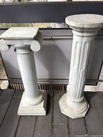 Front view of two classical white concrete pedestals, one square topped with scroll design and one round topped with fluted shaft and octagonal base. Visible minor surface wear present.