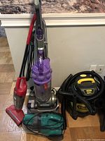 Photo showing four vacuums grouped together: Dyson upright vacuum in purple and silver, red Dirt Devil upright vacuum, green Bissell handheld vacuum, and Stanley shop vacuum with yellow handle and hose coiled around