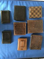 Top-down view of eight men's wallets arranged on blue fabric, showing variety in colors and textures including black, brown, and patterned wallets with bi-fold styles.