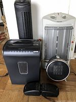 Front view of black tower fan, white heater, black paper shredder, and small black speaker on floor against wall.