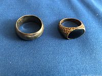Two rings displayed side by side on blue fabric: plain silver band and decorative gold signet ring with black onyx.