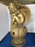Front view of the gold plaster cherub statue showing the cherub sitting on a star-cutout sphere with floral decorations.
