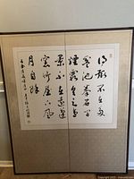 Framed Japanese calligraphy scroll showing black ink characters and red seals on white paper with beige matting and dark frame.