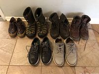 Photo showing six pairs of men's shoes and boots in size 9.5 arranged on tiled floor, featuring various colors and styles including dress shoes and boots made of leather and suede materials.