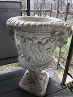 Concrete garden planter with ornate figures and reliefs detailing, standing on sculpted pedestal base.