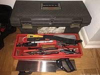 Black plastic toolbox closed with a red tool tray on top containing saws, pliers, screwdrivers, and other hand tools.