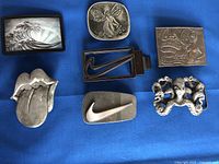 Seven metal belt buckles arranged on a blue surface showing variety of themes including wave, fairy, Coca-Cola, Nike, octopus, and tongue designs.