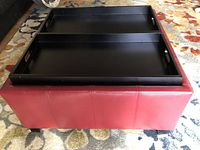 Side angle view showing red leather storage ottoman with two black flip-over trays on top