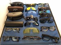 Top-down view of the collection showing all sunglasses, eyeglasses, cases and replacement lenses arranged on a blue surface.