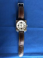 Front full view of the square stainless steel watch with brown leather strap laid flat on blue fabric.