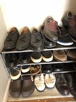 Multiple pairs of men's shoes on metal racks showing variation in style, color, and condition. Shoes include dress shoes, sneakers, casual slip-ons, and clogs. Different materials such as leather, fabric, and synthetic are visible.