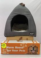 Front view of gray soft, triangular pet house with white plush cushion inside, displayed on top of an orange box labeled 'Warm House for Your Pets' featuring a dog image.