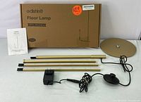 Floor lamp components including cardboard box, manual, three gold rods, circular base, power cord with adapter and black touch dimmer switch.