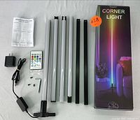 Photo showing disassembled components of the RGB corner floor lamp including multiple LED tubes, two black mounting bars, assembly screws, a remote control, an AC adapter, and instructions laid out on a white surface.