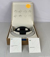 Smart neck massager housed in an open box showing the device with grey ergonomic pads and two user manuals inside the box lid with usage illustrations.