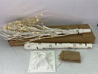 Partially assembled pre-lit white birch tree with warm white LED lights turned on, showing the lighted branches and realistic birch bark trunk sections with base stand and burlap wrap beside it.