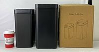 Two black smart trash cans of different sizes beside a coffee cup for size reference. One smaller and one larger bin. Cardboard box labeled 'Smart Trash Can' shown.