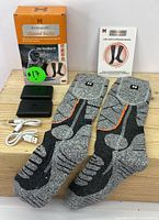 Pair of rechargeable heated socks with packaging, instruction manual, two battery packs, and two charging cables laid out on wood surface.