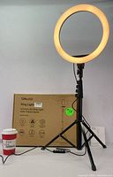 Photo showing the 12-inch LED ring light illuminated with warm light on the adjustable tripod stand, including its cardboard packaging in the background and a coffee cup for size reference.