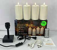 Lot with four white LED flameless candles standing on box, black lamp base, power adapter cord, remote control, USB charging cables and instruction manual laying on table.
