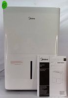 Front view of white Midea 50-pint dehumidifier with water level window and user manual in front.