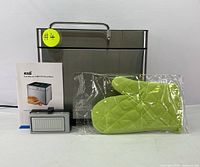 Front view of stainless steel bread maker with handle, user manual, measuring tools, and green oven mitt in packaging.