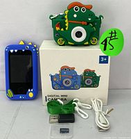 Photo showing green dinosaur-themed kids digital camera, blue touchscreen device standing upright, white USB cables, green wrist strap, 32GB memory card and black reader placed on a white surface with the camera box in the background.