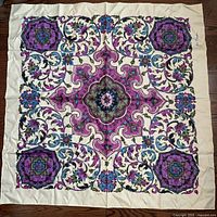 Large vintage square silk scarf with purple, blue, green, black floral and ornamental design on cream background, laid flat on dark wooden floor.