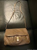 Front view of woven leather purse laid flat on black surface showing tan color, brass pull with paper wrap, and long strap.