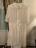 Front view of the white cotton-silk gauzy dress hanging on a hanger, showing full length, shirt collar, gathered waist, and button front from collar to waist.