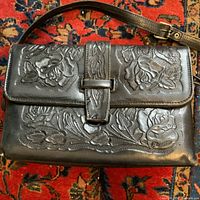 Front view of the black leather handbag showing hand-tooled floral rose patterns on the flap and body, flap strap closure, and adjustable shoulder strap.