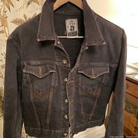 Front view of dark faded jean jacket with button closure and two chest flap pockets showing intentional fraying and contrast stitching.