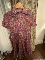 Front view of the full dress hanging on a wooden hanger, showing short sleeves, collared neckline, flowing skirt, and attached belt at waist.