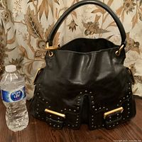 Front view of the black leather handbag with stud detailing, two front buckle pockets, gold-tone hardware, and single top handle next to a water bottle for scale.