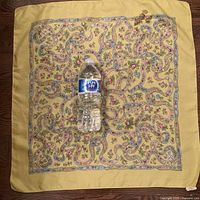 Full view of the vintage silk Liberty of London scarf laid flat with a water bottle for scale, showing a yellow border and winding blue and pink ribbon floral patterns on a lighter yellow background, with visible stain on one corner.