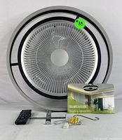 Front view of a circular bladeless modern ceiling fan with built-in LED light, showing its metal grille and black and silver concentric ring design. Included are a black remote control, metal mounting bracket, user manual, and installation hardware components.