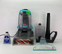 Front view of Bissell carpet cleaner with water tanks, brush rollers visible, accessories and cleaning formula bottle laid out beside it.