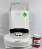 DreameBot D10 Plus robot vacuum placed in front of auto-empty docking station with manual visible behind and a cup for size reference