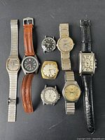 Overview photo showing all seven vintage men's watches arranged in two rows on a dark surface. Varied shapes, strap types, and dial colors visible.