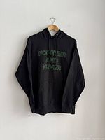 Black men's hoodie hanging on a hanger with phrase 'FOREVER AND NEVER' printed in green outline on the front.
