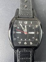 Front view of men's Fossil wristwatch showing black dial with white numerals, day-date window at 3 o'clock, and black leather strap.