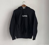 Front view of black Supreme hoodie hung on wooden hanger against white wall, showing front kangaroo pocket and Supreme logo patch.