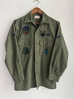 Front view of olive green long sleeve army shirt with sewn patches on chest and sleeve, hanging on wooden hanger.